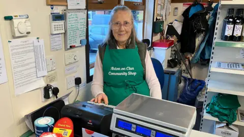 A volunteer standing behind the till smiling into the camera and wearing a green apron. 