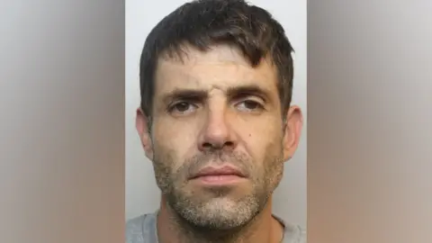 A mugshot of Nehemiah Brown. He has short dark hair, brown eyes and stubble on his chin. He is pictured against a plain background, wearing a light grey t-shirt. He is looking directly at the camera with a blank expression. 