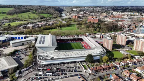 A drone photo of Ashton Gate, taken from a very high angle. It shows crowds going into the stadium, which is in the middle of a built-up area. The River Avon and Clifton Suspension Bridge are visible in the background.