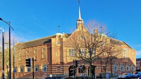 Ian Hughes A large brown-brick building with a small spire above a large entranceway and trees standing outside