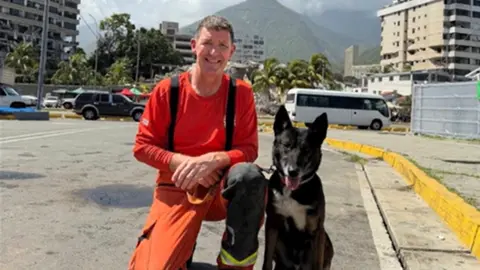 Lancashiire Fire and Rescue Serivice A male dog handler smiles as he poses next to his sitting black sniffer dog