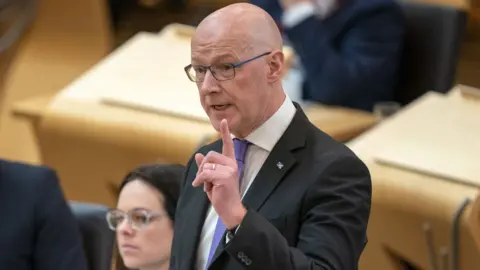 John Swinney, who has a bald head and glasses, speaks in the Scottish Parliament chamber. He is standing with his left hand raised in front of his body, with his index finger pointing upwards. He is wearing a dark suit, white shirt and purple tie. 