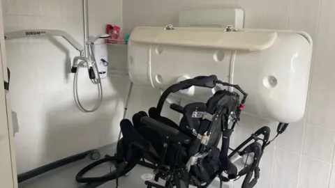 BBC News A wet room with shower, hoist and changing table. The tiles are white and the floor grey.