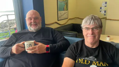 Two men sit on a blue sofa. The walls behind them are cream coloured with a painting of a beach hanging on it. The man on the left wears a long sleeved black t-shirt and wears a red and black watch. He has a bald head, grey beard and holds a white mug. The man on the right Has short grey hair, wears clear glasses and a black t-shirt.