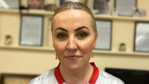 A close-up image of Tricia O'Neill who is a young woman with blonde hair, which is tied back. She is standing in a light coloured room with some framed images behind her, but they are blurred. She is wearing a white shirt with a red collar.
