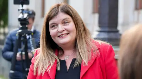 Sioned Williams, who is wearing a red blazer and black blouse. She has blonde hair and is wearing a necklace. A man with a camera on a tripod can be seen behind her.
