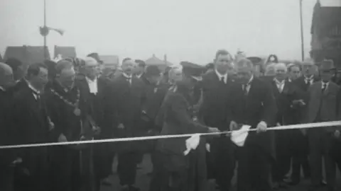 British Pathe A still from a black and white newsreel film of the Prince of Wales in military uniform cutting a ribbon, accompanied by other men in formal dress, uniform and mayoral regalia.  