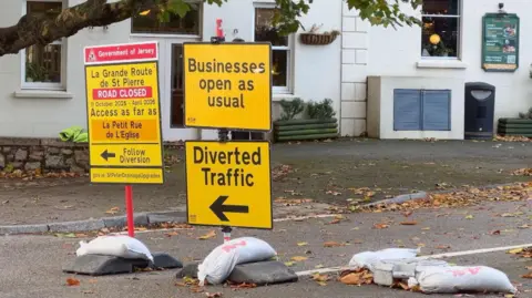 St Peter's Main Road is blocked by diversion signs being held down by sandbags outside the George de Carteret pub. One sign reads Businesses open as usual. The others give signage for diverted traffic to follow.