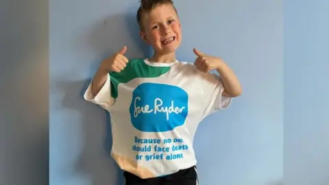 Isaac smiles at the camera while wearing a T-shirt with the Sue Ryder logo and making thumbs up sign