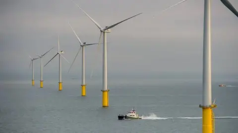 Rampion Offshore Wind Six white wind turbines with yellow bases in a row in the sea. There is a small boat in between the first two in the image.