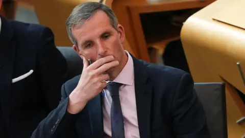 Liam McArthur sitting in the parliament during the debate on his bill. He looks thoughtful and his right hand is over his mouth.
