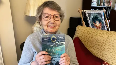 Margaret Imrie sitting on a sofa holding a copy of her book on the history of Hull's Lit and Phil. She has grey hair and wears glasses and is wearing a grey top.