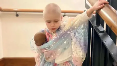 Handout A girl who has a bald head following chemotherapy treatment is wrapped in a shawl made from sparkly silver material and is wearing a white top. She is holding a doll under her right arm and he right hand is resting on a wooden banister rail which is held up by black iron railings