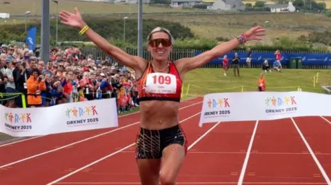 Christa Cain raises her arms as she crosses the finish line. She wears a red and black lycra running top and shorts, and sunglasses. She runs towards a white finishing line ribbon that has just split in two either side of her. A full crowd watches on.