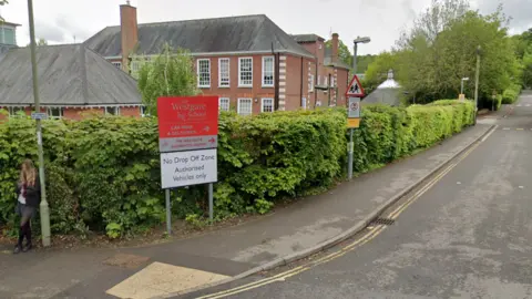Google A red school sign saying 'The Westgate School'. The sign is next to a hedge and on the corner of a road. There is a brick building behind the hedge.