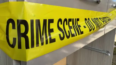 A chest of drawers stands in a street with yellow crime scene tape around it.