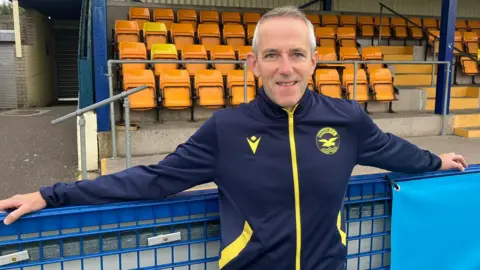 Stephen McCullough, con cabello corto y una sonrisa, se encuentra cerca de una balaustrada marina que lleva una parte superior en una pista amarilla y azul. Tiene los brazos estirados a lo largo de la balaustrada mientras se inclina allí.