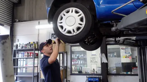 A man in a blue cap and shirt looking at the front wheels of a blue car, which is raised up