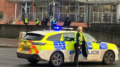 A police officer stands beside a police car while other officers talk to pedistrians