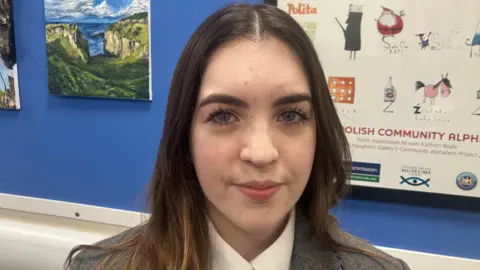 Niamh is smiling at the camera. She has long dark hair and is wearing a grey blazer and white shirt. She is standing in front of a blue notice board. The board has a painting of seaside cliffs.