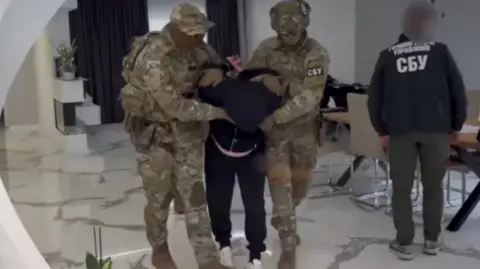 SSU Ukraine said a Hungarian spy was detained in the eastern Ukrainian region of Transcarpathia. A man dressed in black can be seen in between two men dressed in army camouflage outfits as he is lead out of an apartment