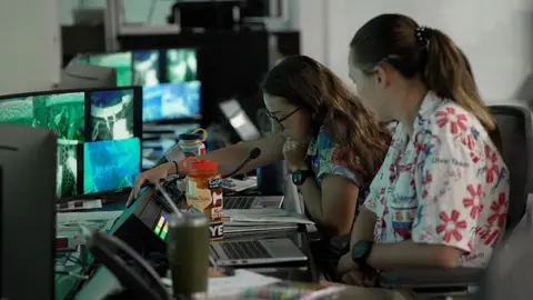 Kevin Church/BBC Trainers  in the control room are leaning in to a microphone and communicating with the astronauts. There is a bank of screens in front of them. The woman on the left is wearing a blue flowery shirt and has brown hair and glasses. The woman on the right is wearing a white shirt with red stylised flowers. The dive is taking place on " wear a Hawaiian shirt to work day."  