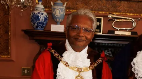 Wearing his mayoral robes and gold chain, Bharat Pankhania smiles at the camera. Behind him two vases and a musical instrument can be seen on a mantelpiece.