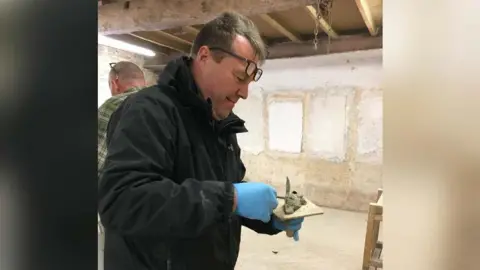 Christine Tallon Adrian Cole putting some cement on a slab. He is wearing a black jacket, blue gloves and has glasses propped on his forehead.