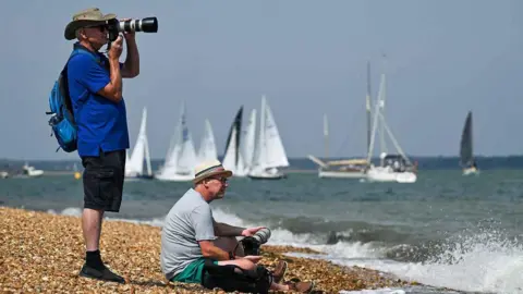 Cowes Week Martin Allen Two people with cameras on a pebble beach watching sailing boats on the water, with waves breaking on the shoreline.