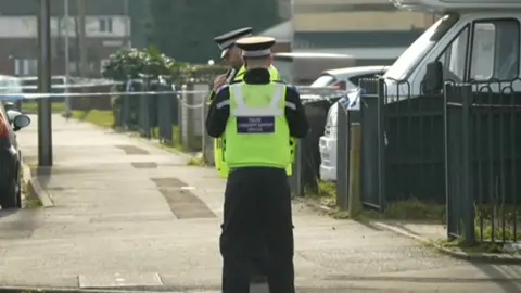 Two police officers stand at the scene with houses and vehicles behind them.