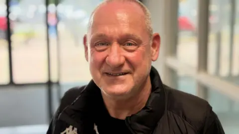 Kitman Roy Dickinson with short grey hair wearing a Wigan Warriors black puffer jacket inside the club's Robin Park Arena. He is smiling.