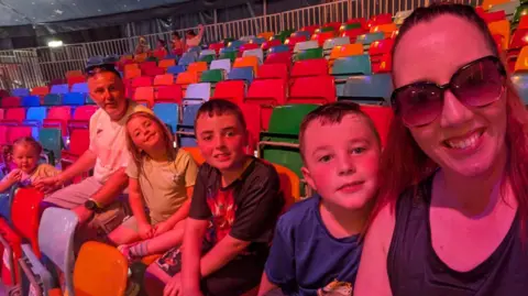 Craig Hinde Craig and Trish Hinde sitting with their four children in rows of brightly coloured seats.