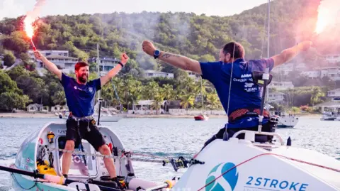 2Stroke.team Karl Austen and Ed Shaw wearing navy blue t-shirts and are holding flares whilst standing on there rowing boat.