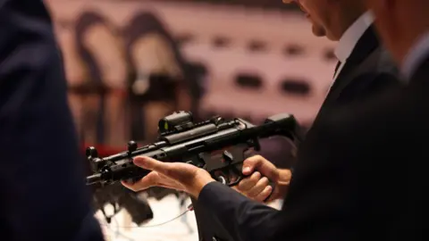 Bloomberg via Getty Images A man in a black suit, his face not visible, holds a black submachine gun in his hands. 