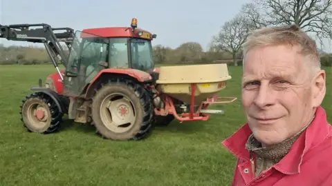 Richard Cornock on his farm in Gloucestershire. He is pictured in a selfie with a red tractor behind him. He is looking at the camera and smiling. 
