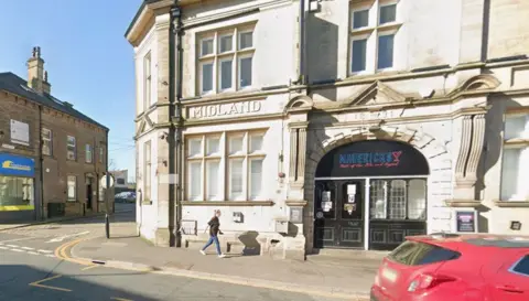 A street corner with a building in the foreground and other smaller buildings in the background. The main building is constructed from light-colored stone and features ornate architecture. Above the central windows the word MIDLAND is engraved in capital letters. Below that there is a large arched entrance, above which is an inscription reading 1874. The entrance has black double doors with glass panels and signage for MAVERICK'S. In the right foreground is a red hatchback car.