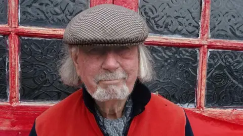 Shane Lucas, a man with a red jumper and a tweed farmer's cap, he has white long hair and a beard and smiles.