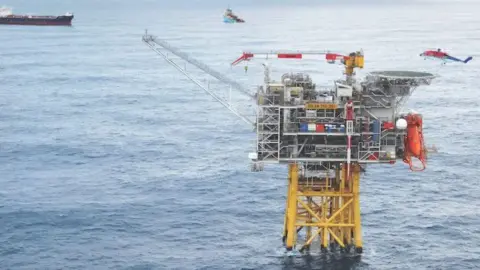 Oil and gas platform with yellow legs in the sea, a blue, red and white offshore helicopter hovers overhead, and vessels in the background. 