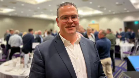 A man wearing a suit in a room of people, background blurred.