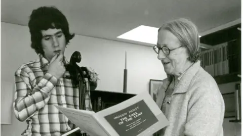 Britten Pears Arts Imogen Holst readers a music sheet as Steven Isserlis stands next to her also looking at the sheet music. Imogen wears glasses and a cardigan while Steven has dark curled hair and wears a chequered shirt with a cello next to him. The image is in black and white.