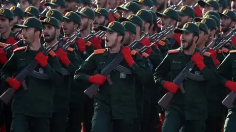 ABEDIN TAHERKENAREH/EPA-EFE/REX/Shutterstock Iranian Revolutionary Guard Corps (IRGC) soldiers march in formation in September 2024. The group of men wear matching green and red uniforms and hold rifles 