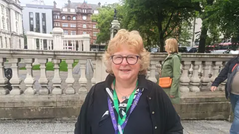 A woman with reddish grey hair and black framed glasses stands in the grounds of Belfast City Hall. She is wearing a black cardigan and shirt