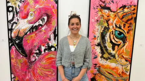 BBC Woman smiling with brown hair standing in front of two brightly coloured paintings on a white wall