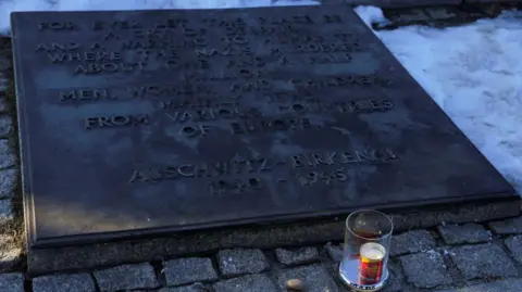 Reuters A candle in a clear jar burns beside a plaque marking the liberation of the Nazi's largest concentration camp at Auschwitz-Birkenau. it is surrounded by snow.