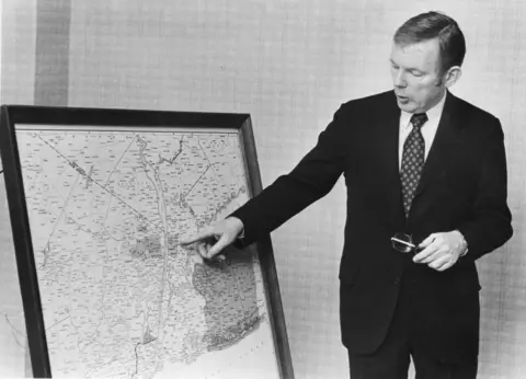 Black and white photo of Charles Dolan in a dark suit pointing to a map of New York