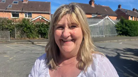 A woman, with long blonde hair, wearing a white top and smiling at the camera.