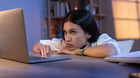 A forlorn young woman slumped over a desk looking at a laptop
