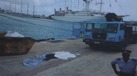 supplied by Chris Purkess A pile of clothes lies on a quayside near a skip and a heavy truck. A cruise ship is in the background and a uniformed officer stands near the camera-taker.