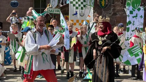 A man dressed in white and green with red trousers at a street party surrounded by knights and jesters. A woman wears a crown and medals.
