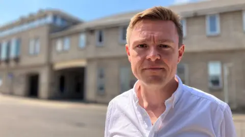 Lewis Adams/BBC Will Quince wearing a white unbuttoned shirt on a sunny day. He has light brown hair and is looking at the camera with a slight frown.